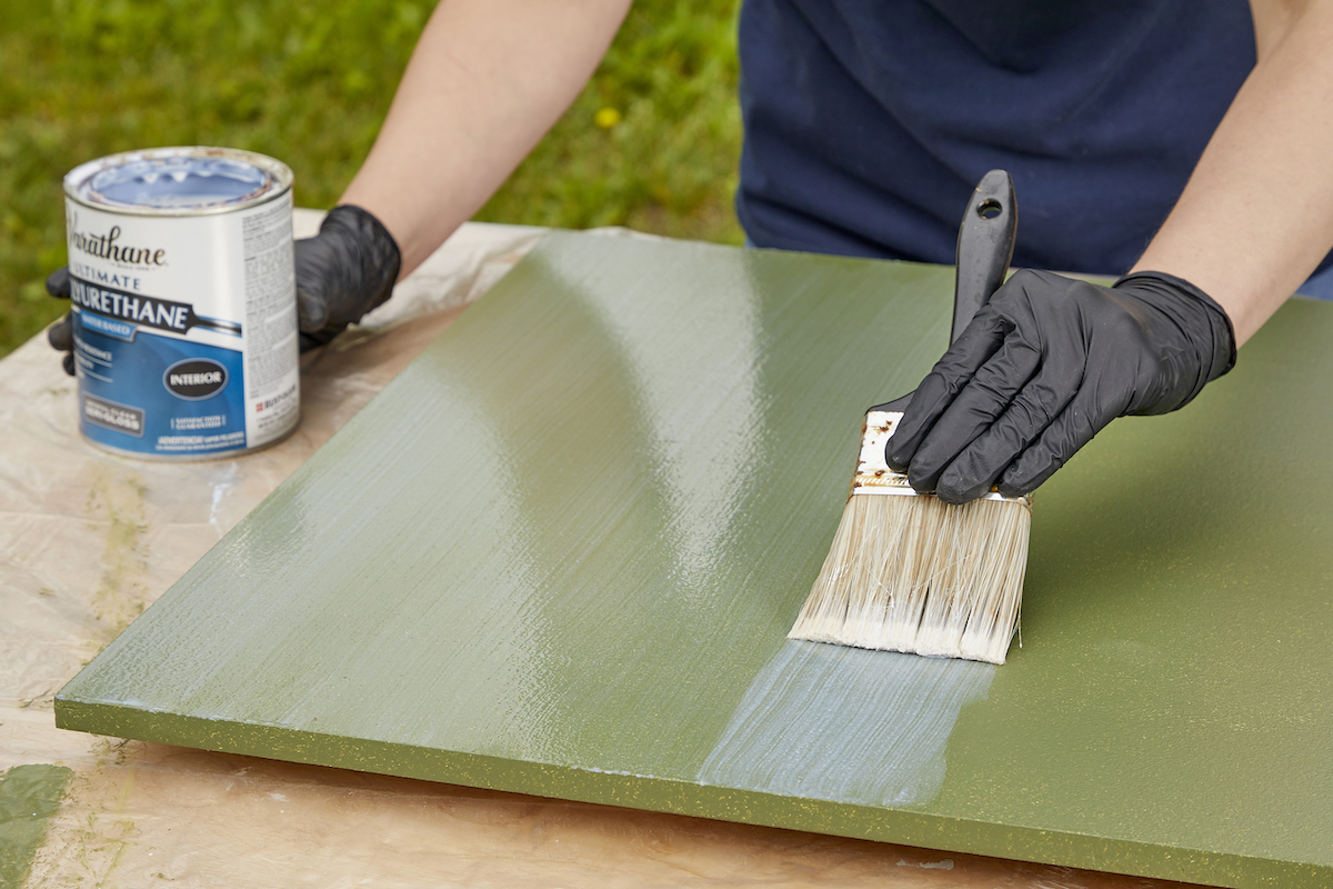 Woman uses a paint brush to apply a coat of polyurethane to a piece of MDF that's painted green.