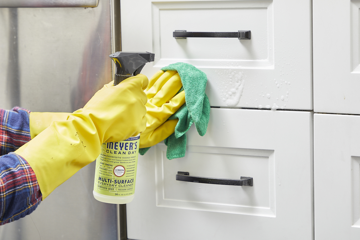 Woman wearing yellow rubber gloves sprays cabinets with cleaner and wipes with a cloth.