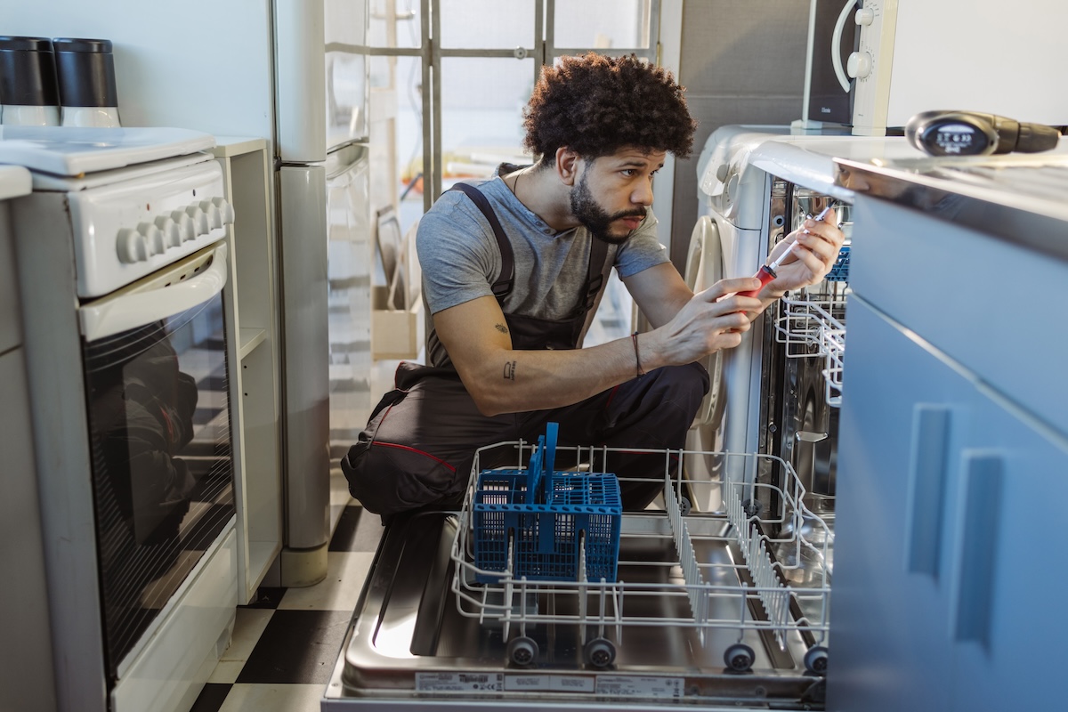 Younger man uses tools to examine where dishwasher fan is in kitchen with blue cabinets. 