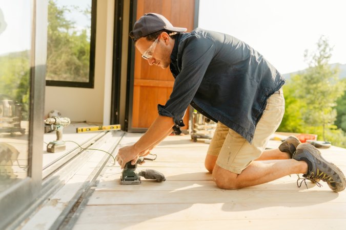 A carpenter with eye protection and grey hat uses a sander on outdoor decking.