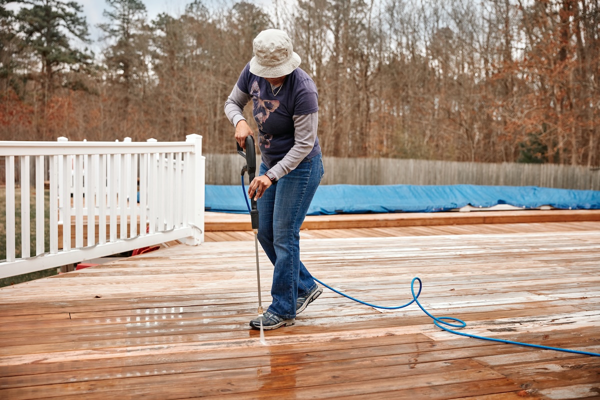 A woman wearing a bucket hat power washes a wooden deck on a cloudy day. 