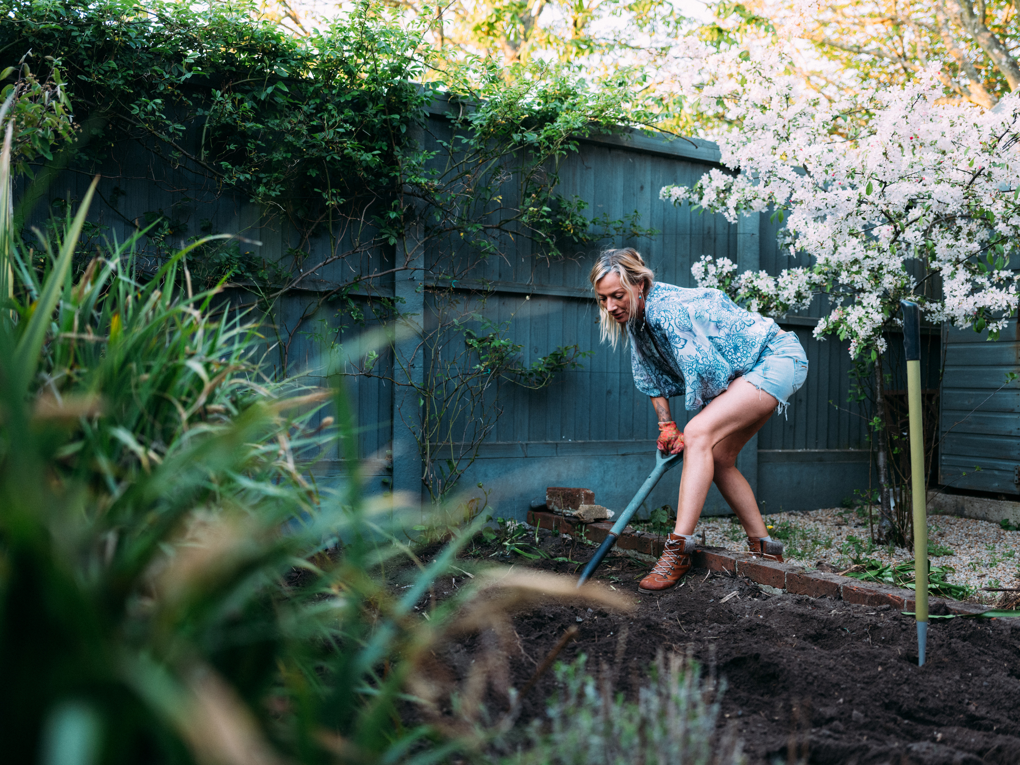 A mature woman wearing leather boots and gloves is digging up a large garden bed in her backyard.