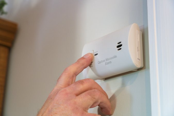 A person is pressing the test button on a carbon monoxide detector on a house wall.