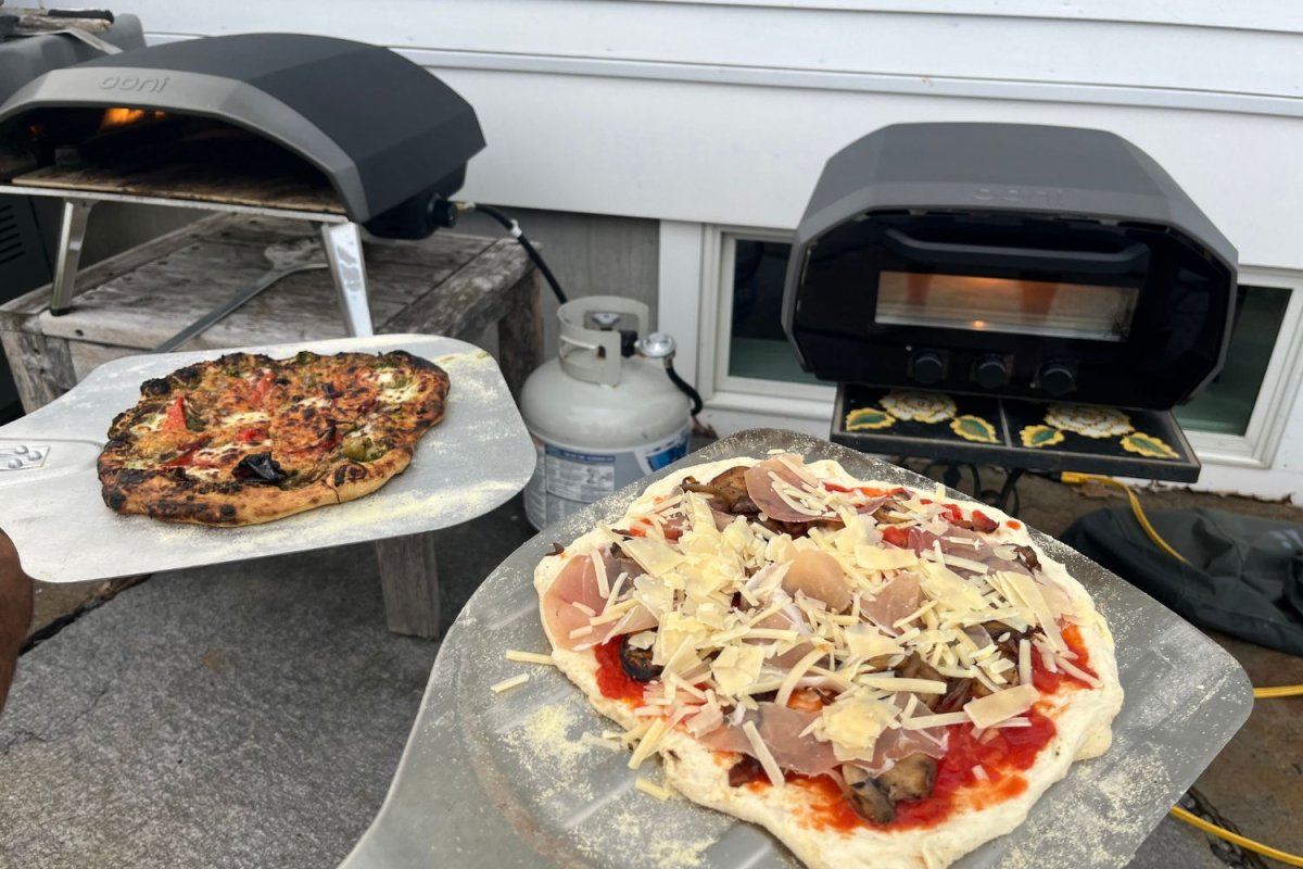 One cooked pizza and one uncooked pizza in front of two outdoor pizza ovens