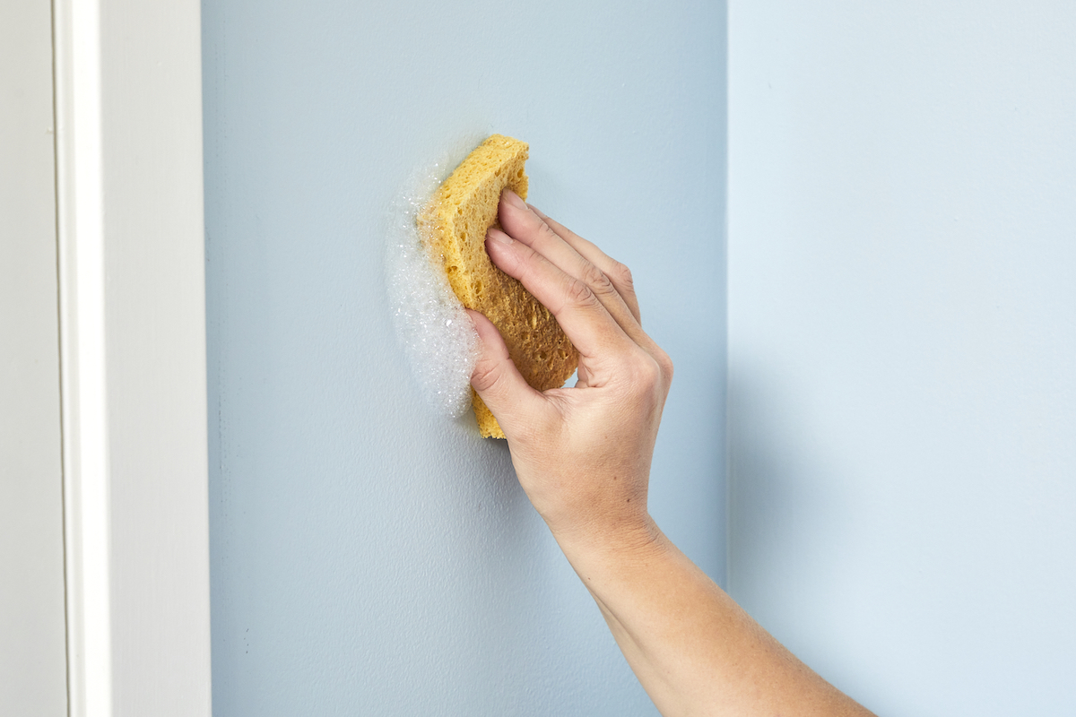 Woman wipes a blue wall with a yellow, soapy sponge.