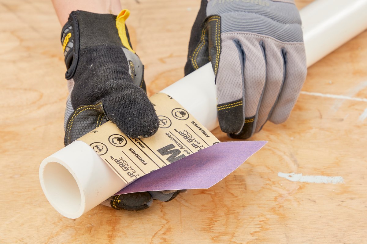 Woman wearing work gloves uses sandpaper to sand a white PVC pipe.