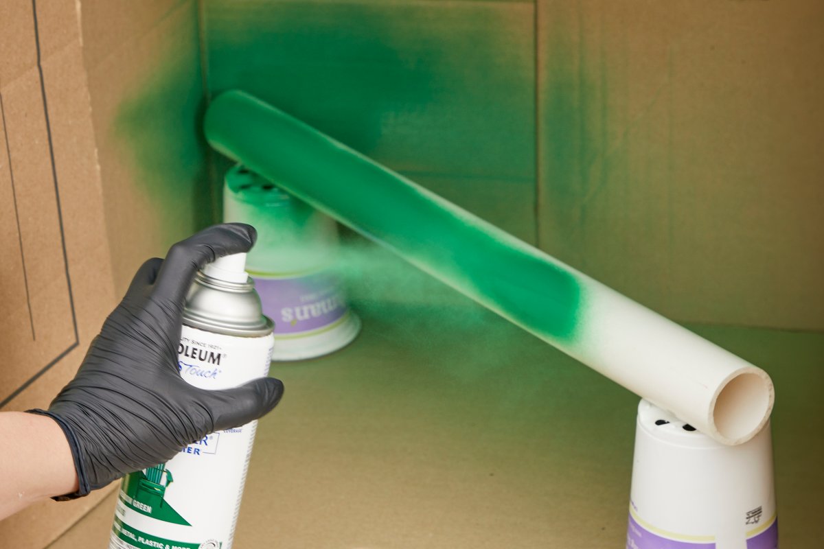 Woman sprays green spray paint on a PVC pipe propped inside a cardboard box.