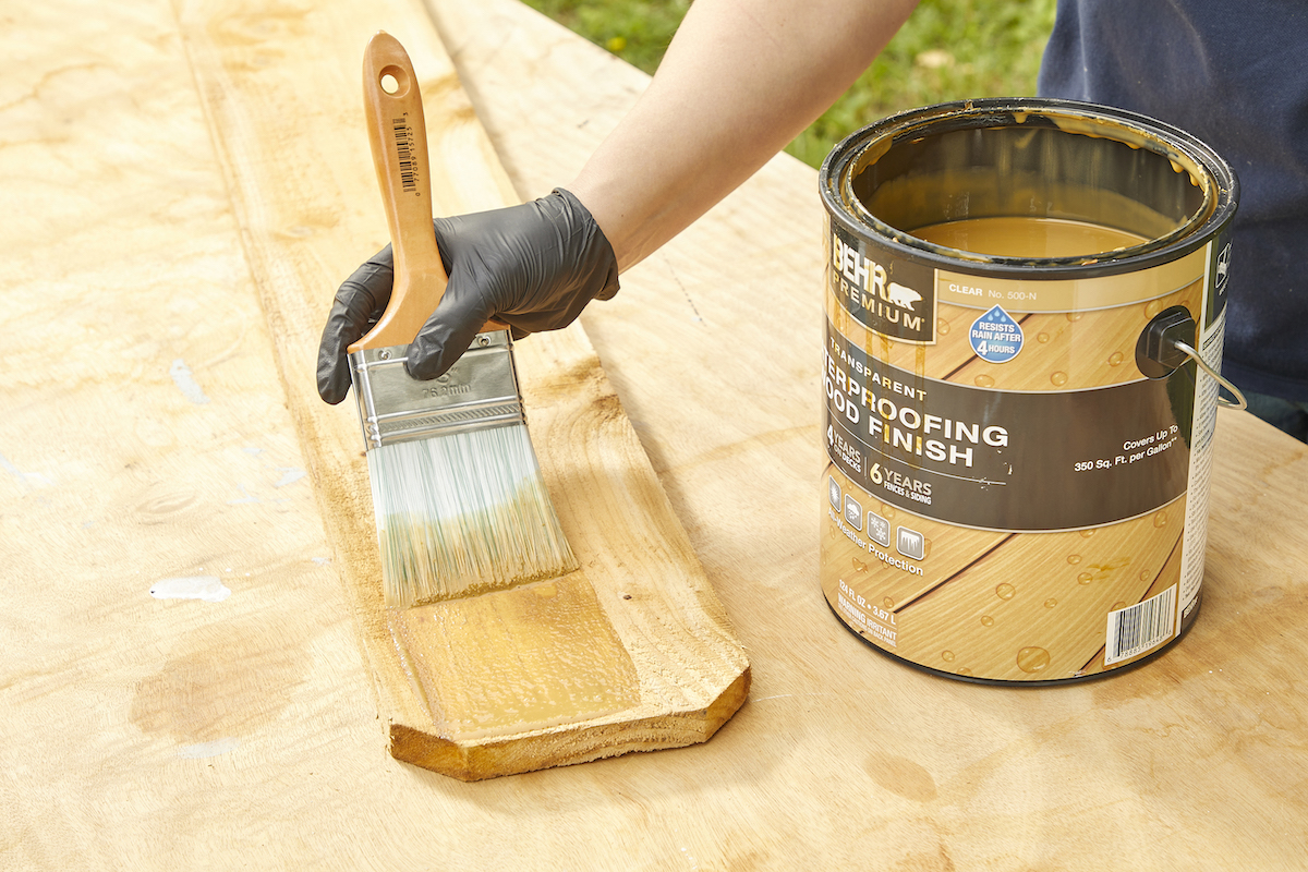 Woman uses a wide paint brush to stain a 2x4 of pressure-treated wood.