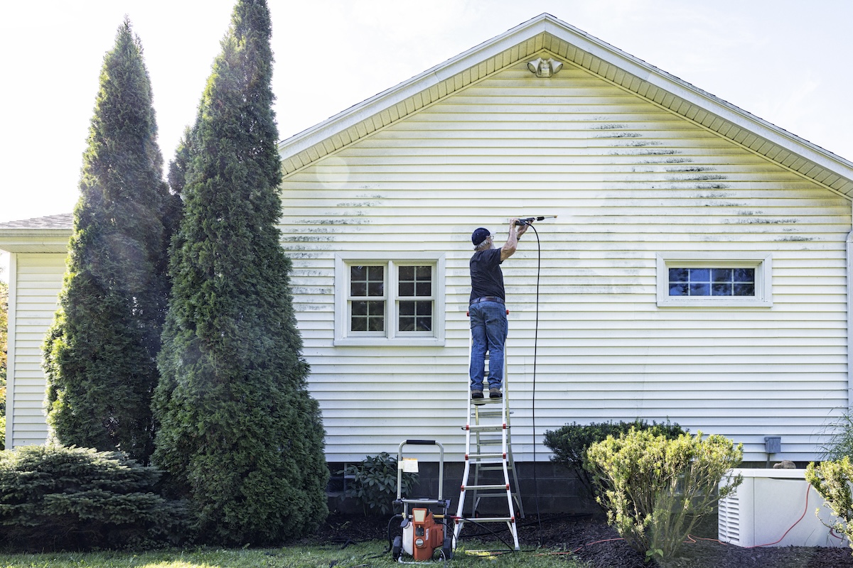 Mature man stands on ladder and uses pressure washer to clean house siding.