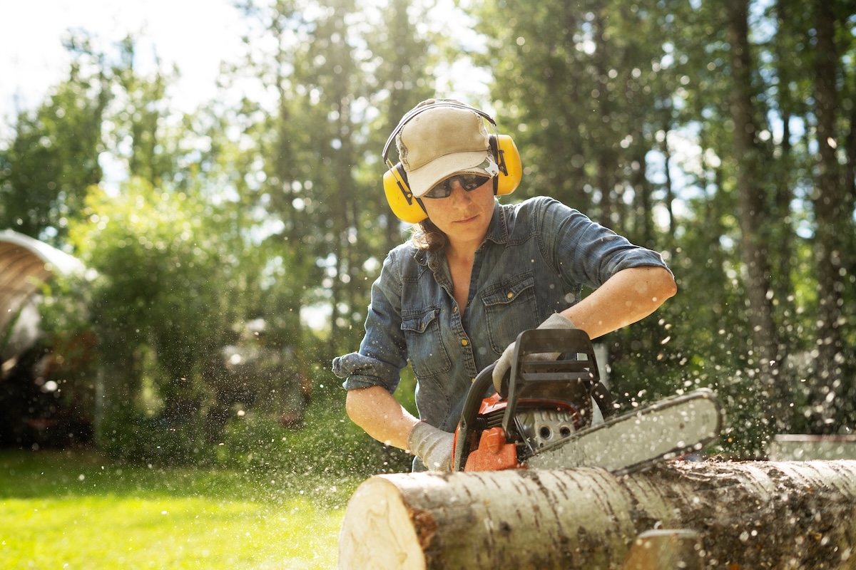 A women wearing ear and eye protection uses a chainsaw on a log.