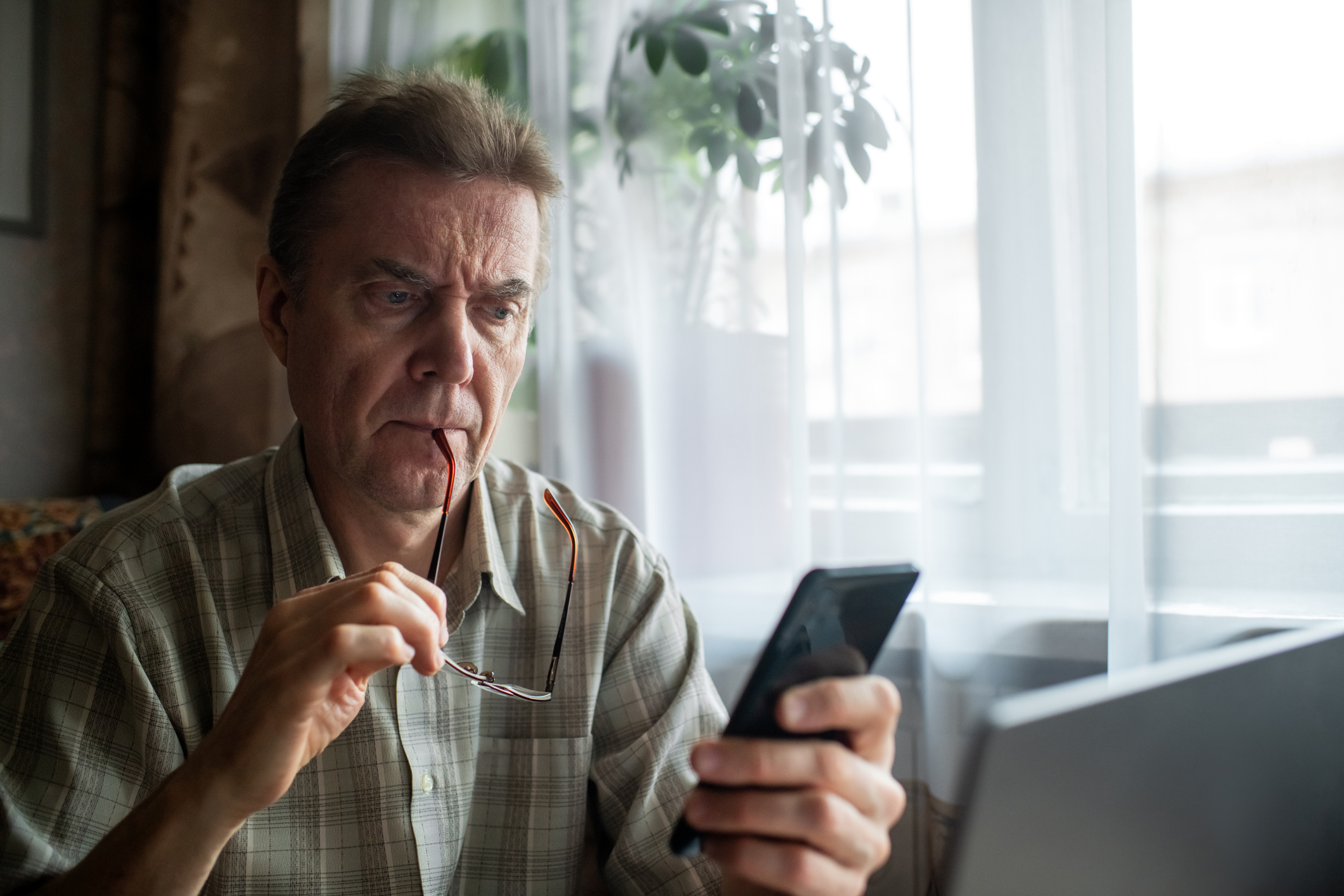 A concerned-looking senior man looks seriously at his smartphone while sitting in front of a laptop.