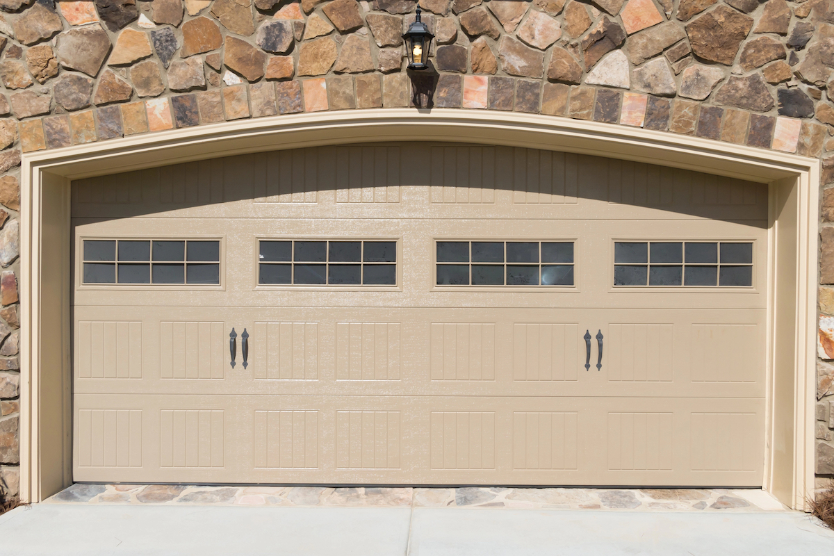 A house with a stone exterior has a beige painted steel garage door with small windows across it.