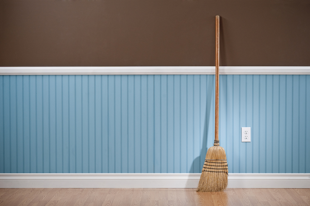 A wooden broom is standing on hardwood flooring and is leaned against a blue and brown wall.