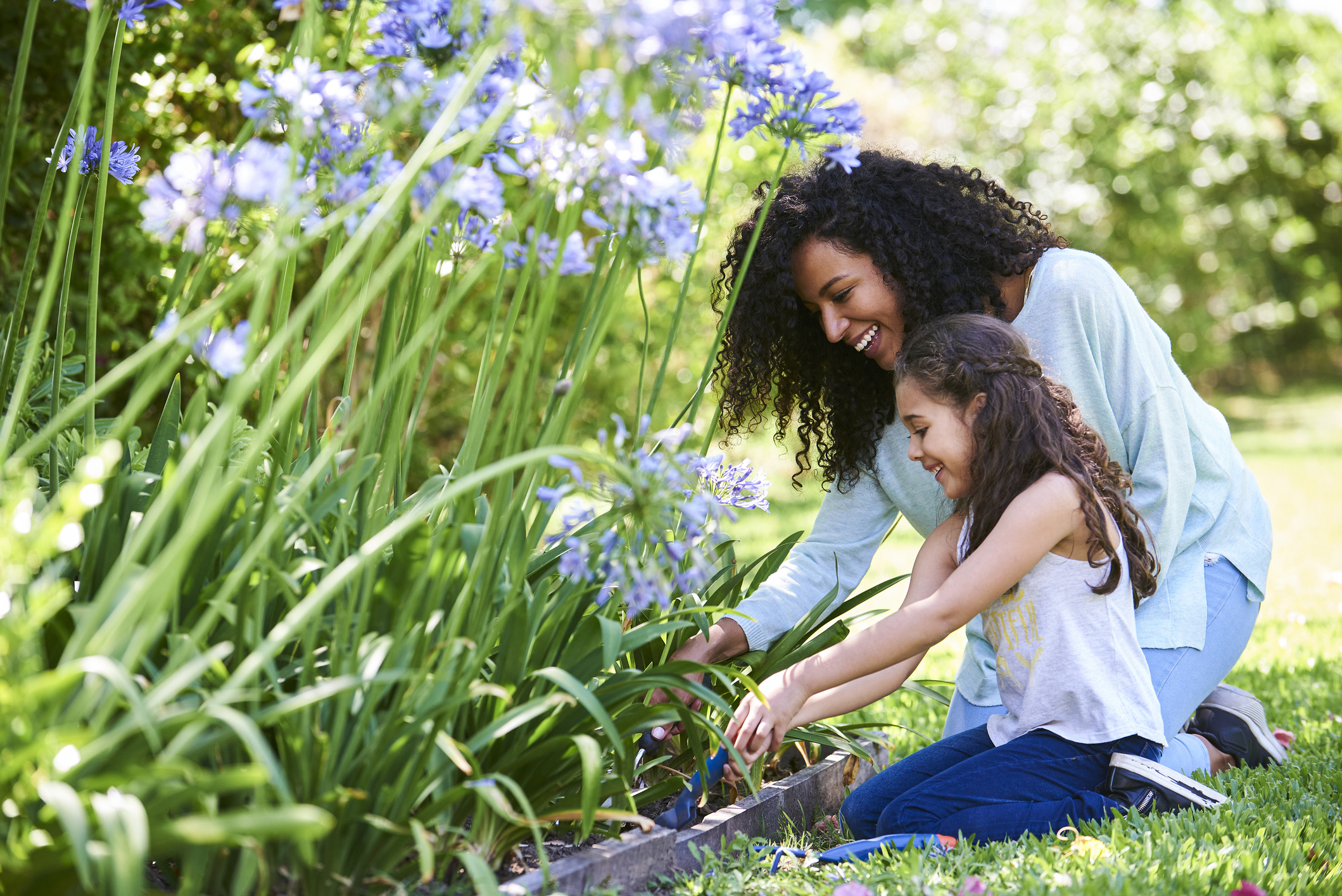 Smiling mother and daughter gardening outdoors by purple flowers.