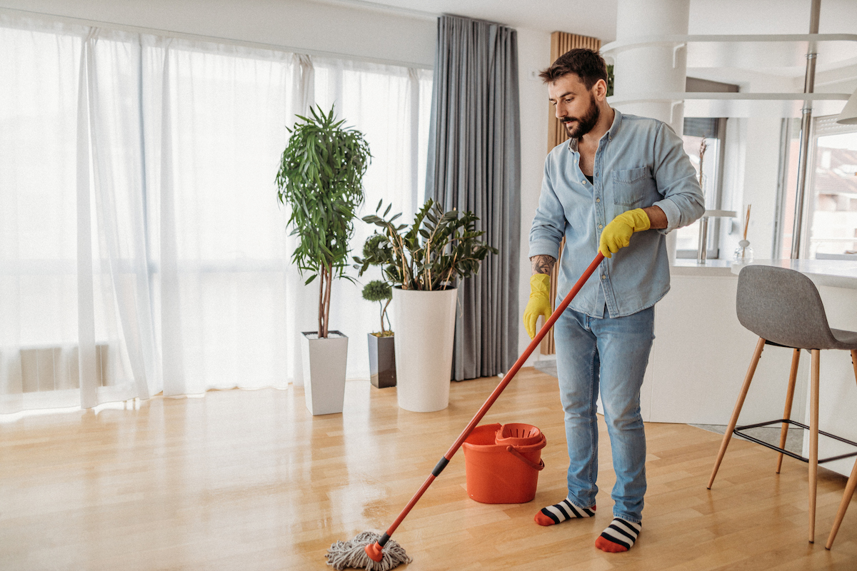 A man wearing yellow gloves is mopping wet hardwood flooring in front of a large window.