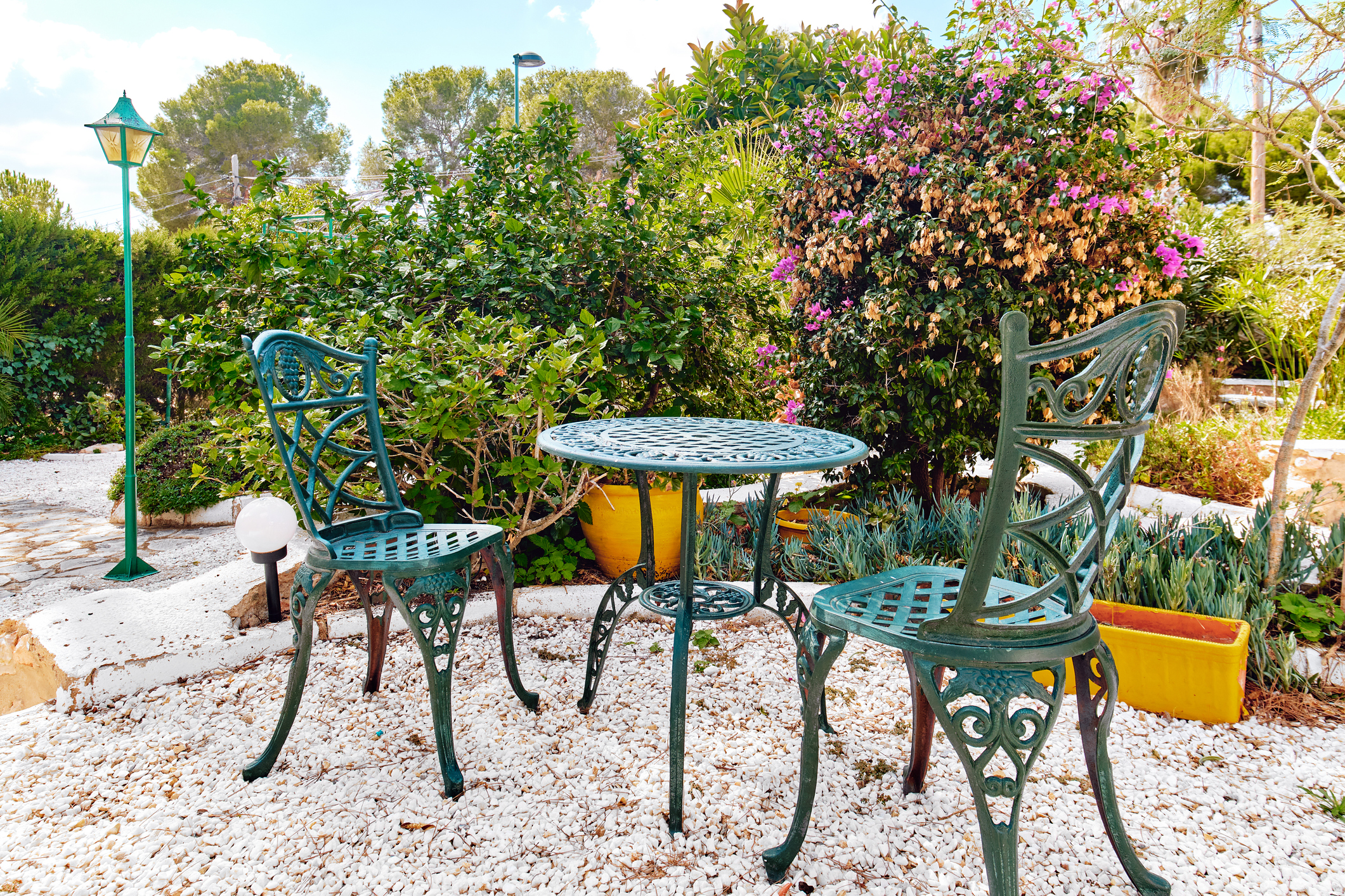 Teal colored metal chairs and table on outdoors patio with blooming flowers in background.