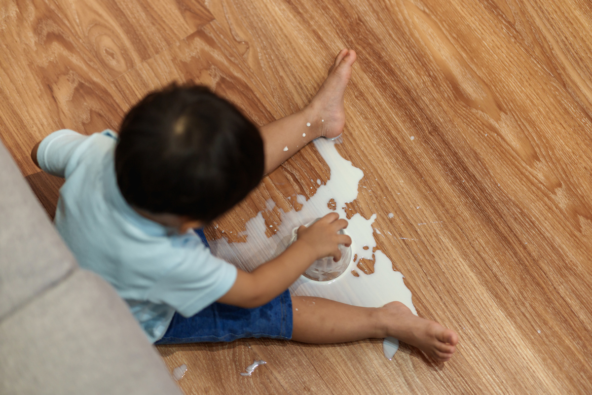 A child is sitting on hardwood floor with an overturned bowl in hand and spilled milk across the floor.