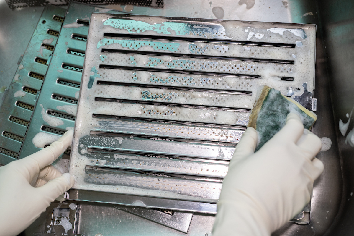 A person wearing gloves is using a sponge to clean a range hood's filters in a sink.