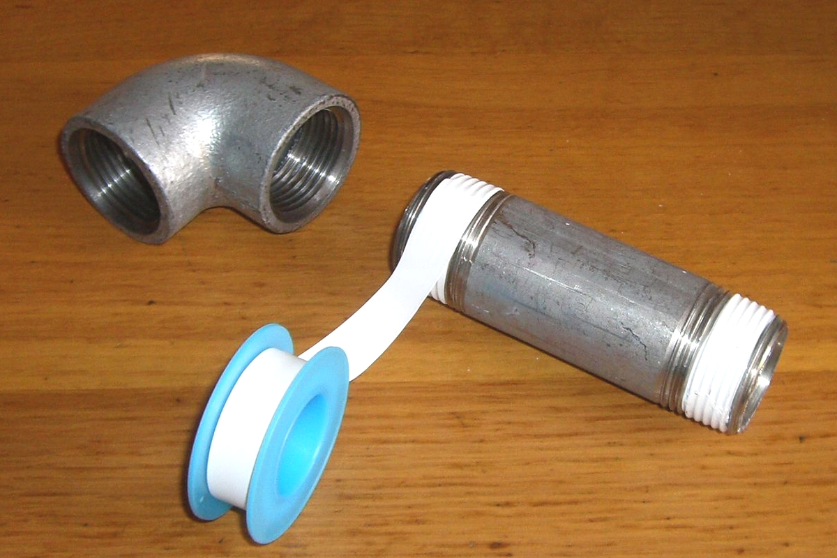 Half-applied plumber's tape on a blue spool, the large pipe to which it is attached, and a connector lie on a wooden table.