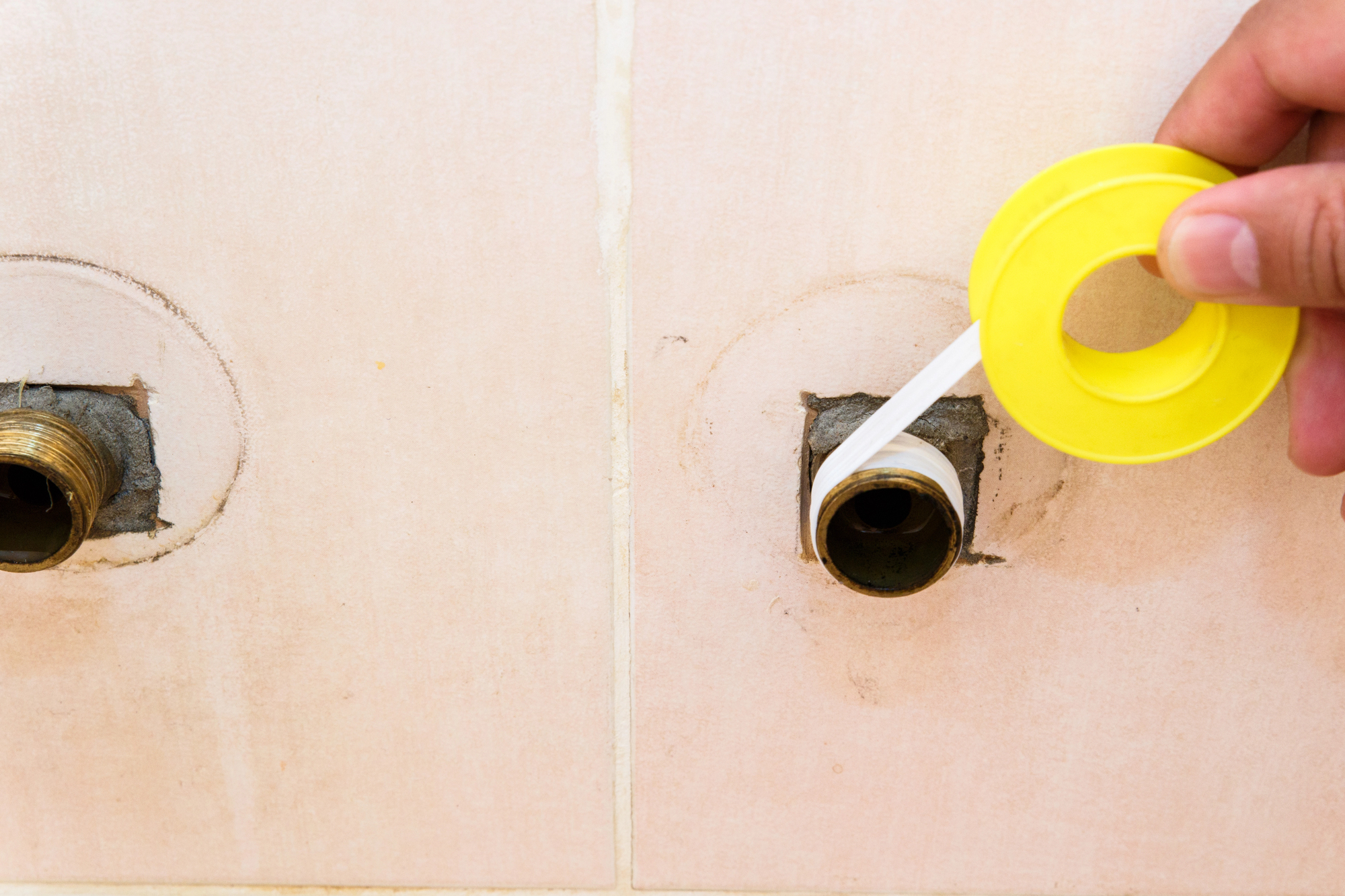 A person holds a yellow roll of plumbing tape taut while sealing the threads of a water supply pipe before installing a fixture or valve on it.