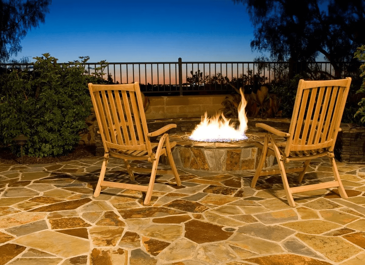 A pair of wooden chairs surround a stone outdoor fire pit.