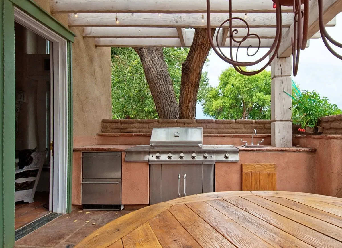 An outdoor kitchen features a stainless steel grill, sink, and drawers and wooden table.