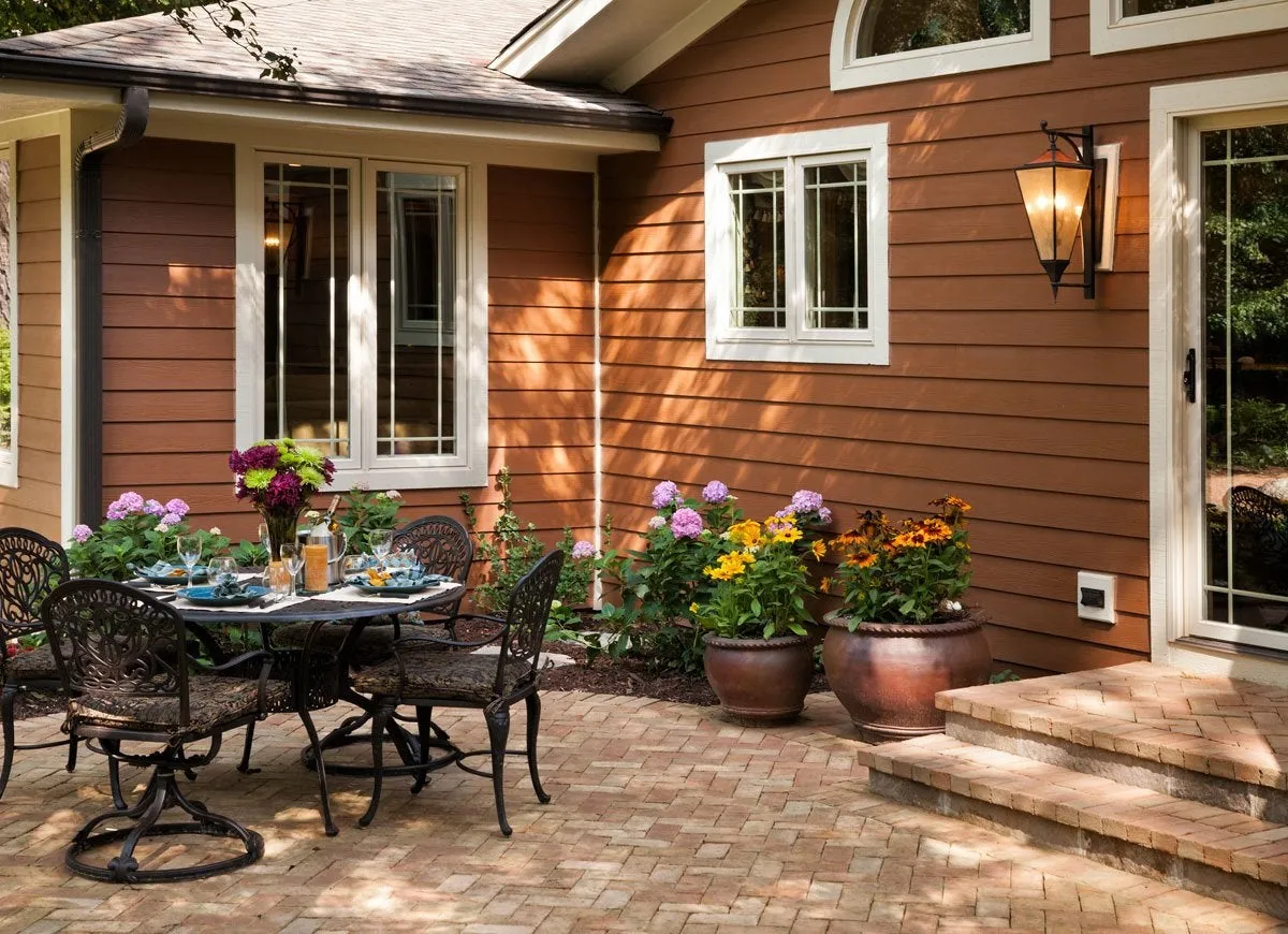 A stone backyard patio features black metal table and chairs and colorful flowers in large pots.