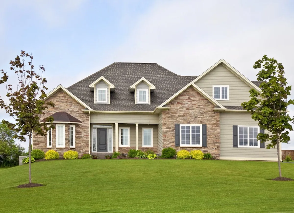 A home with manufactured stone veneer and vinyl siding has a large lawn and two young trees.