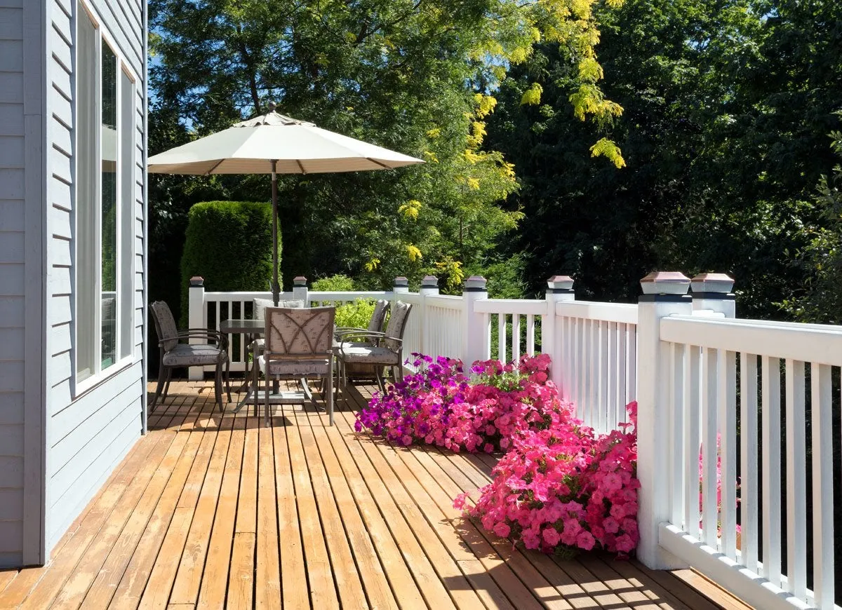 A wooden deck is lined with white railing and decorated with an outdoor table set and pink flowers.