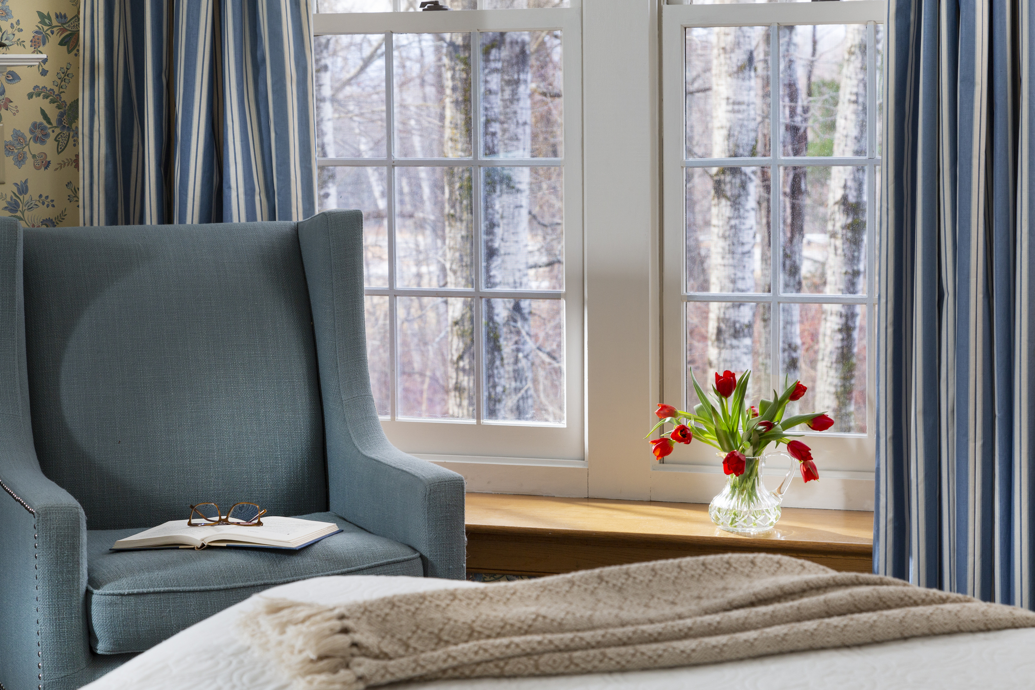 A spare bedroom window with open blue-and-white striped curtains, a blue reading chair, and made bed.