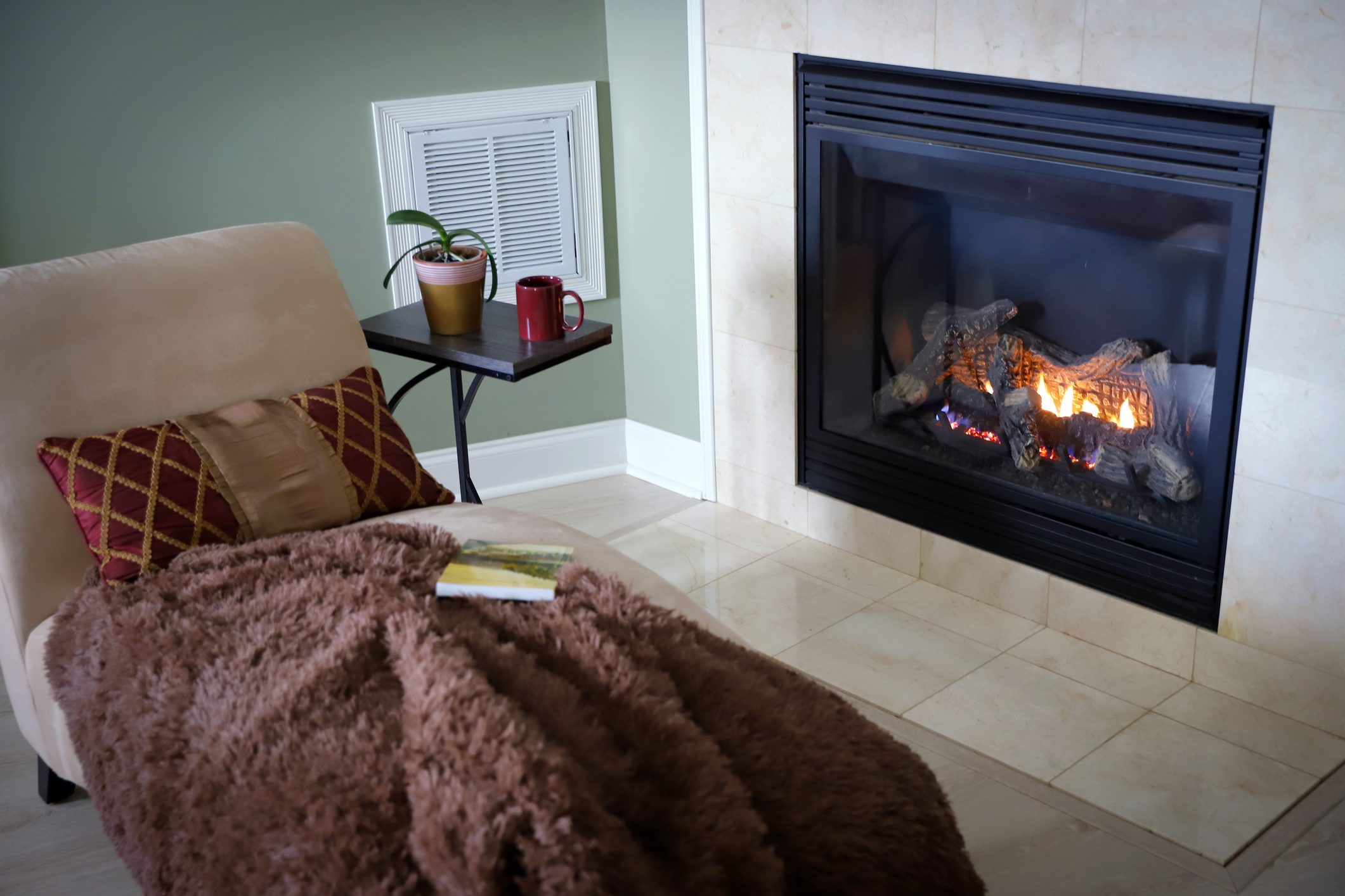 A cozy beige chair with a blanket, pillow, and book on the seat is placed in front of an electric fireplace insert.