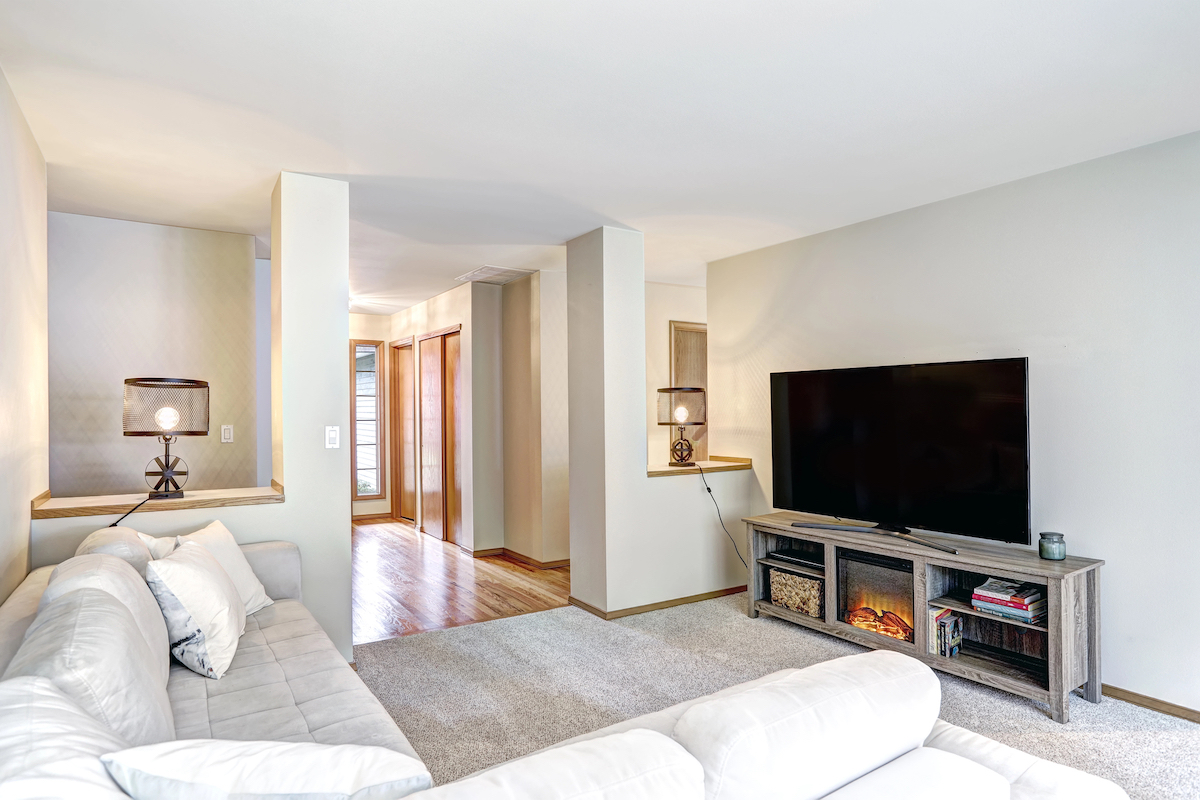 An electric fireplace stand is supporting a TV in a living room with a white couch and white rug.