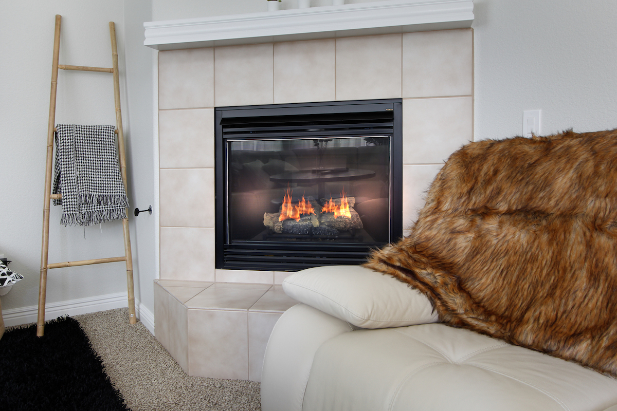 A black direct-vent gas fireplace is installed in white stone in a living room with a white couch covered by a fur blanket and a ladder holding a blanket. 