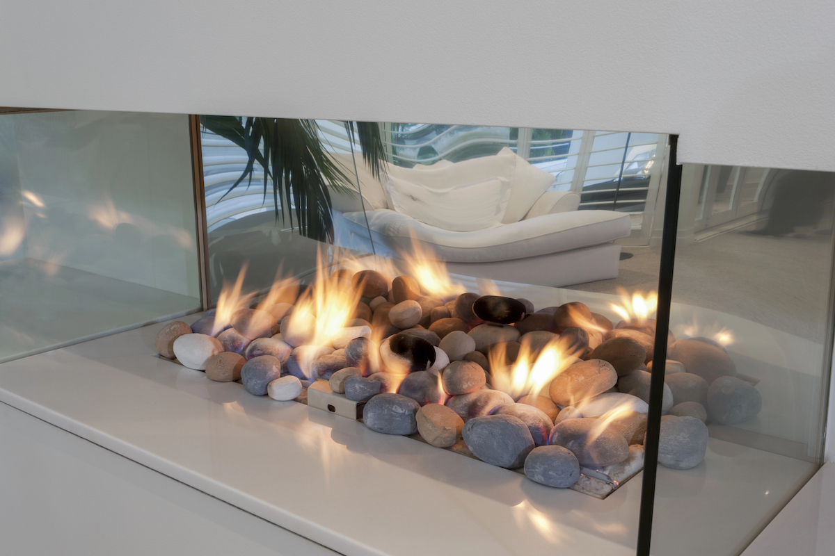 A fireplace with stones burning in encased in glass, allowing a view of the white chair in the next room.