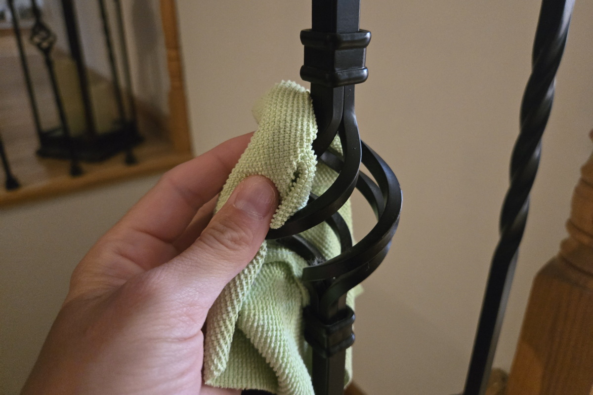 A woman wipes down the ornate spindles of a wrought iron staircase with a light-green microfiber cloth.