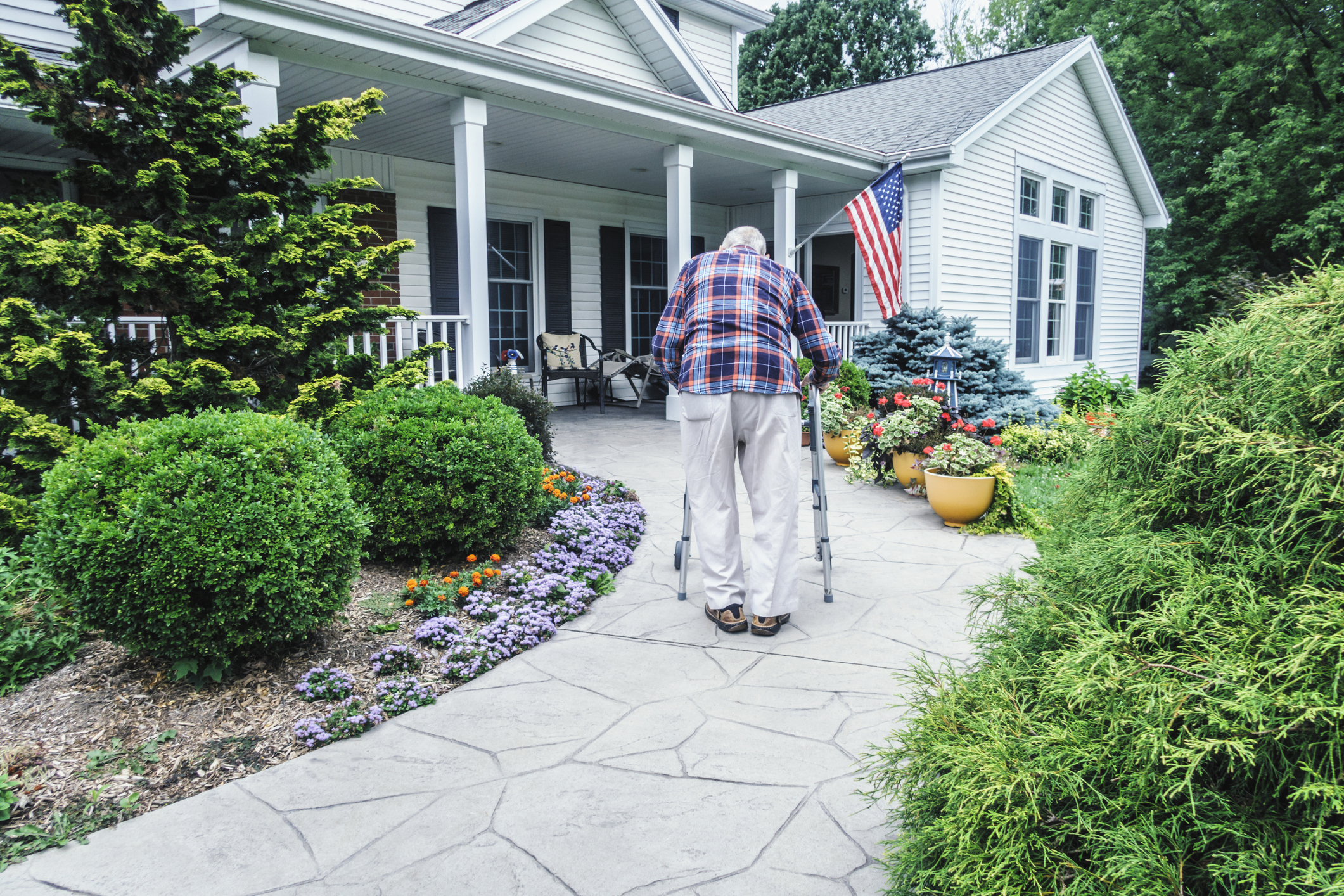 An elderly man using a walker to walk up to a house with a ramp to the front door.