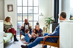 Grandmother sitting on floor with two granddaughters, while their parents and grandfather sit by them in chairs and watch.