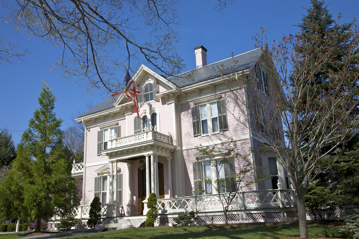 A pink colonial style home with an American flag attached to the front is shadowed by large trees.