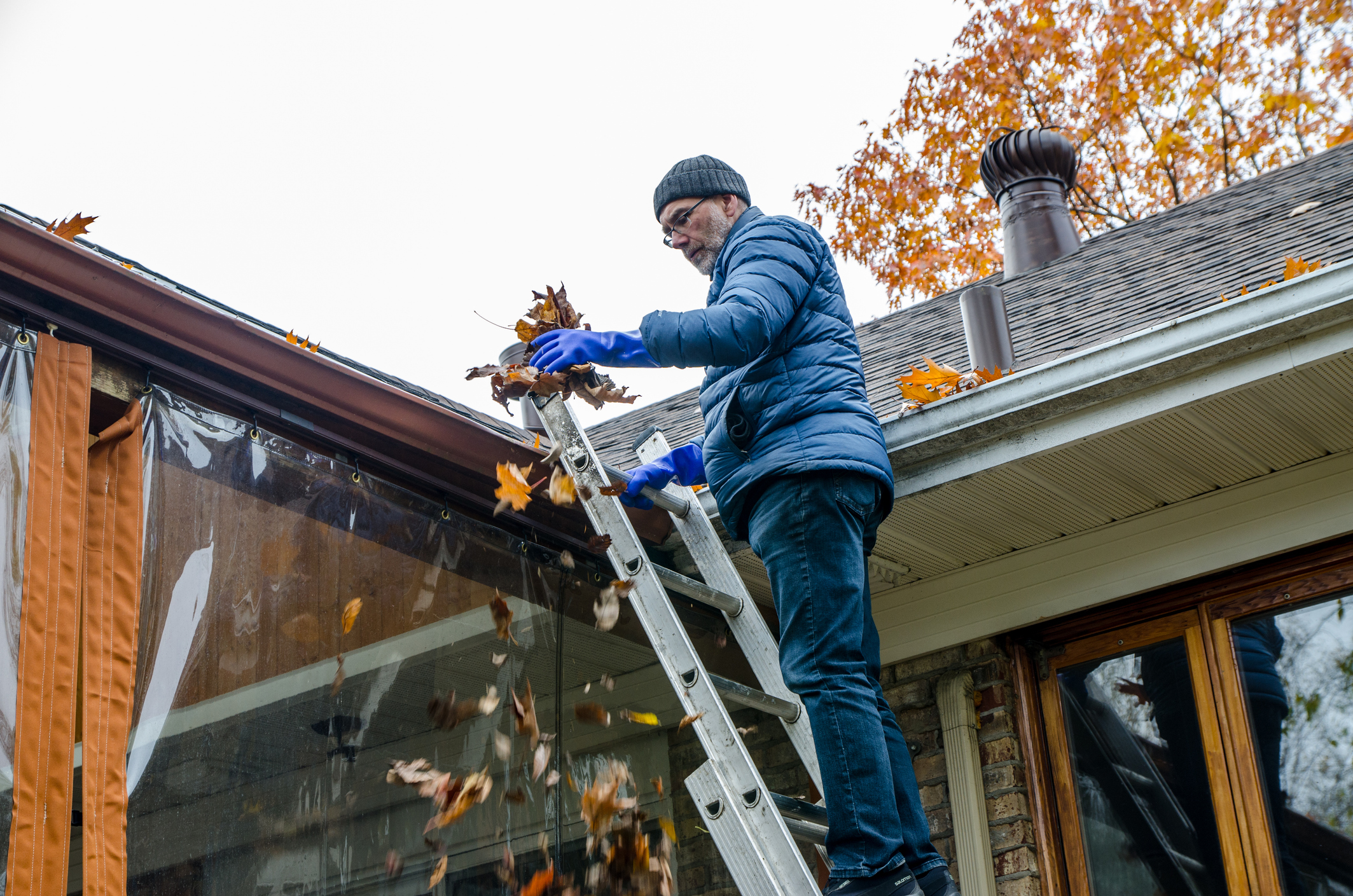 Man on ladder removing autumn leaves from gutter on his house.