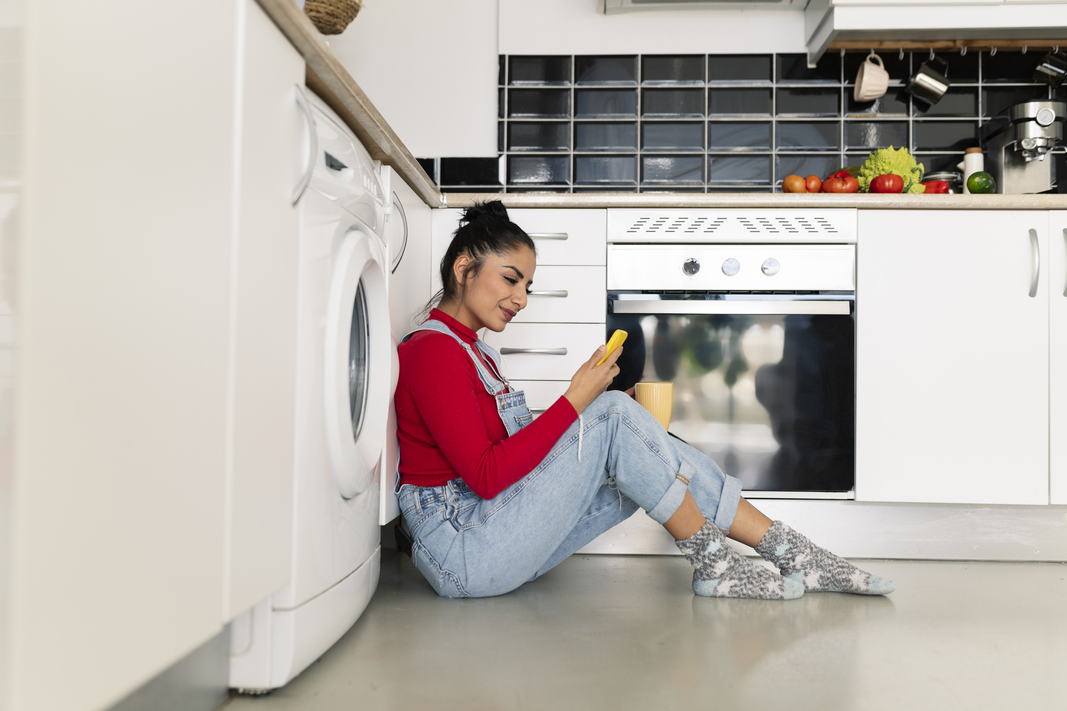 Woman sitting on the kitchen floor in front of a washing machine waiting for the laundry to get done.