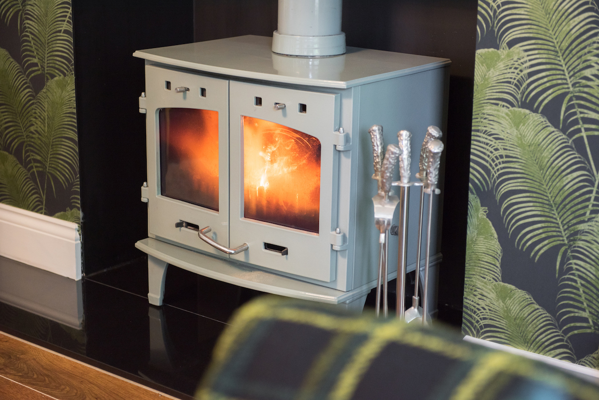 A light grey wood burning stove is burning wood in a room with tropical leaf wallpaper.