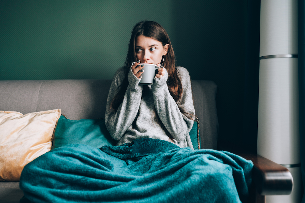 A nervous-looking woman is sitting on a grey couch wearing a sweater with a teal blanket on her lap while drinking from a mug.