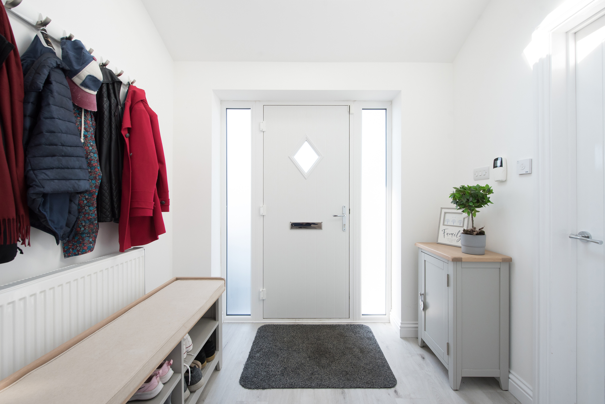 Interior view of a one-story foyer and hallway painted white, with front door, doormat, grey console cabinet, potted plant, coat rack with coats, shoe rack bench.