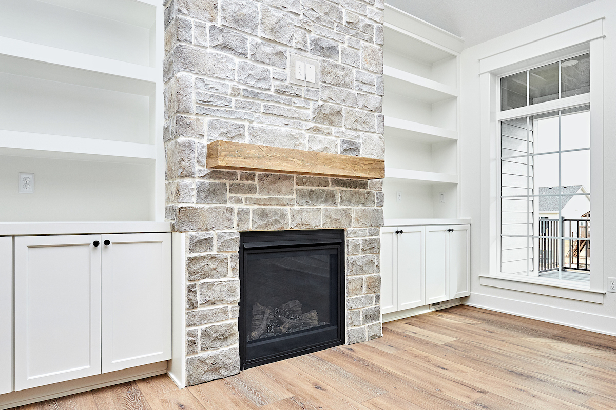 A B-vent gas fireplace is installed in white stone inside of a living room with empty white built-in shelving.