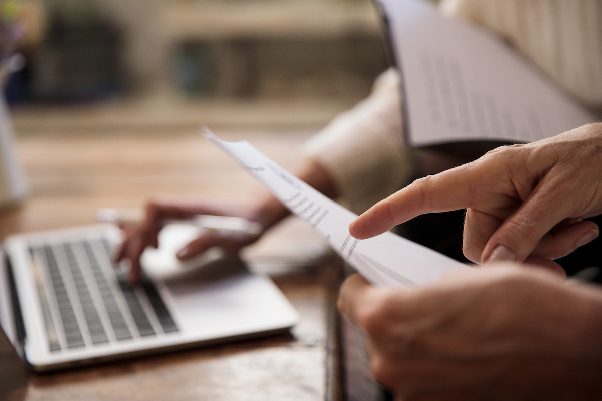 Two people are reviewing printed documents while one of them is using a laptop on a table.