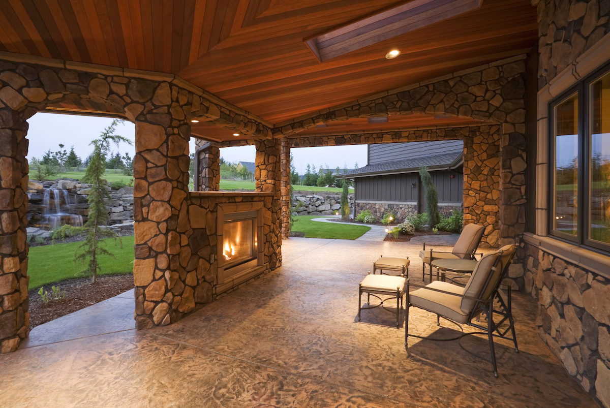 A covered backyard patio features a stone fireplace fueled by stone, chairs, and small tables.