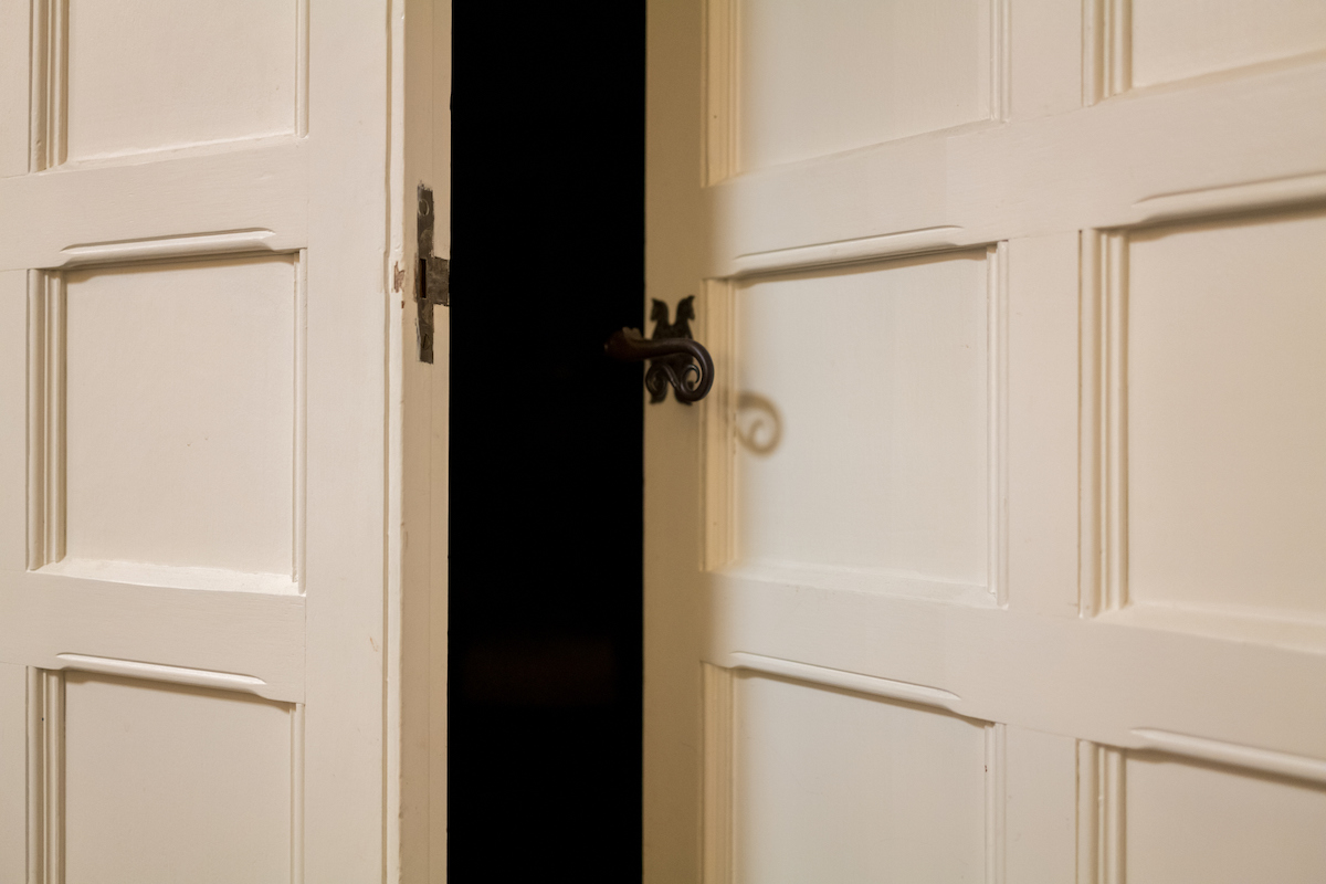 A white wooden door with a metal latch is ajar to view a darkened room behind it.