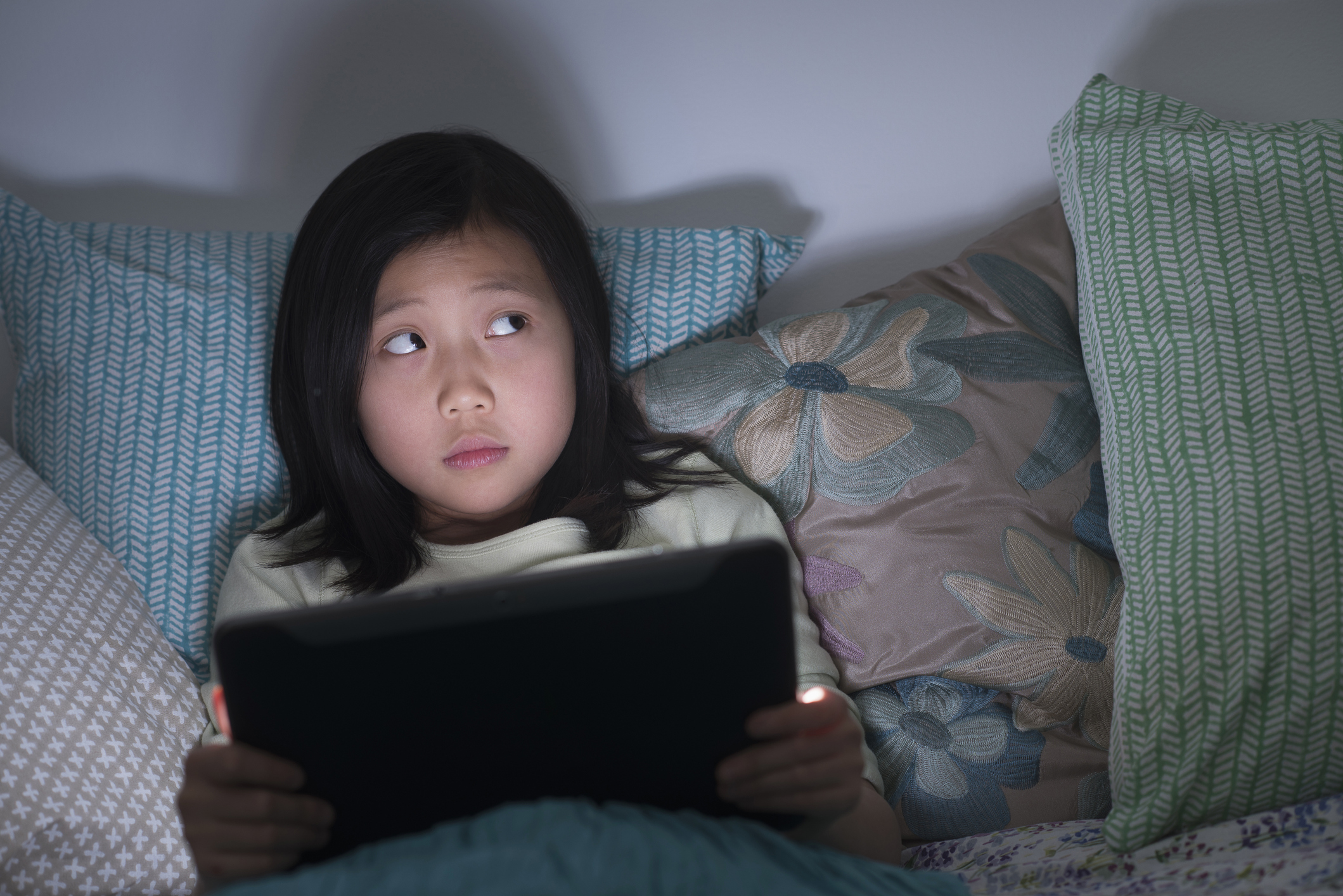 A girl who's holding a tablet in bed looks up at the ceiling with a scared expression.