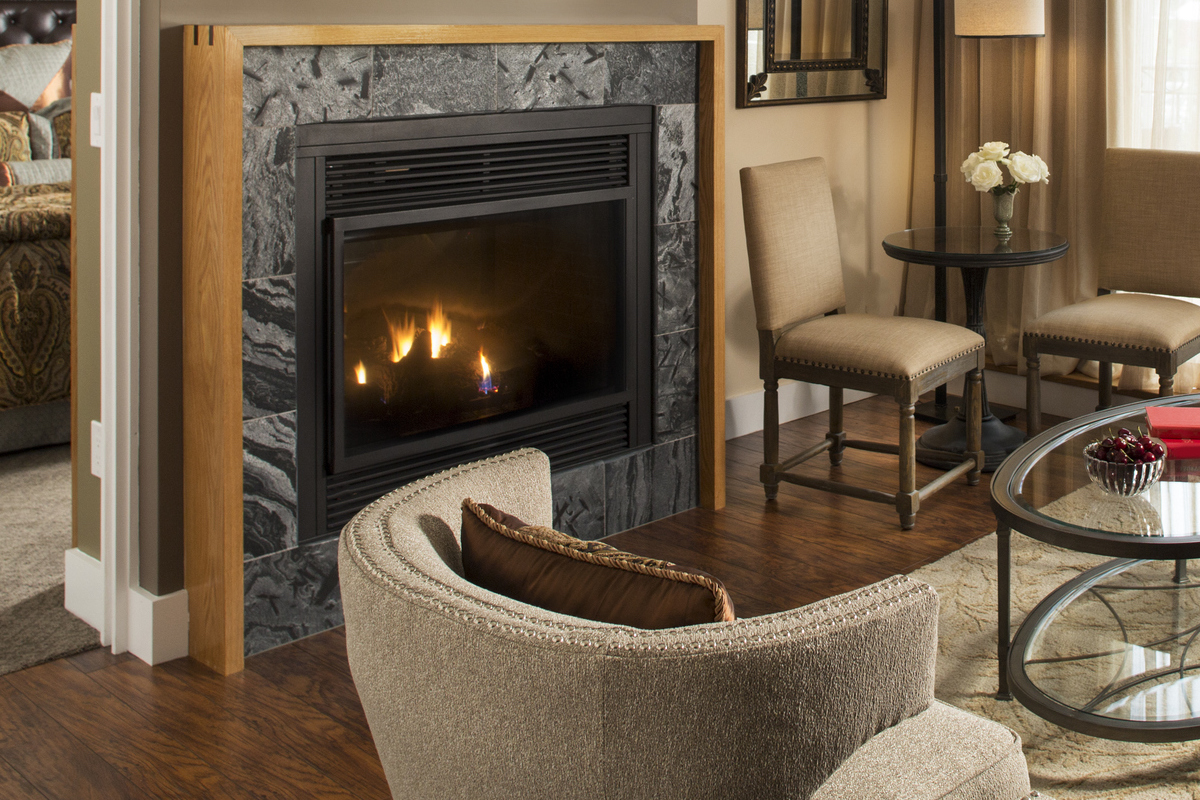 A ventless gas fireplace burns in a living room furnished with taupe chairs.