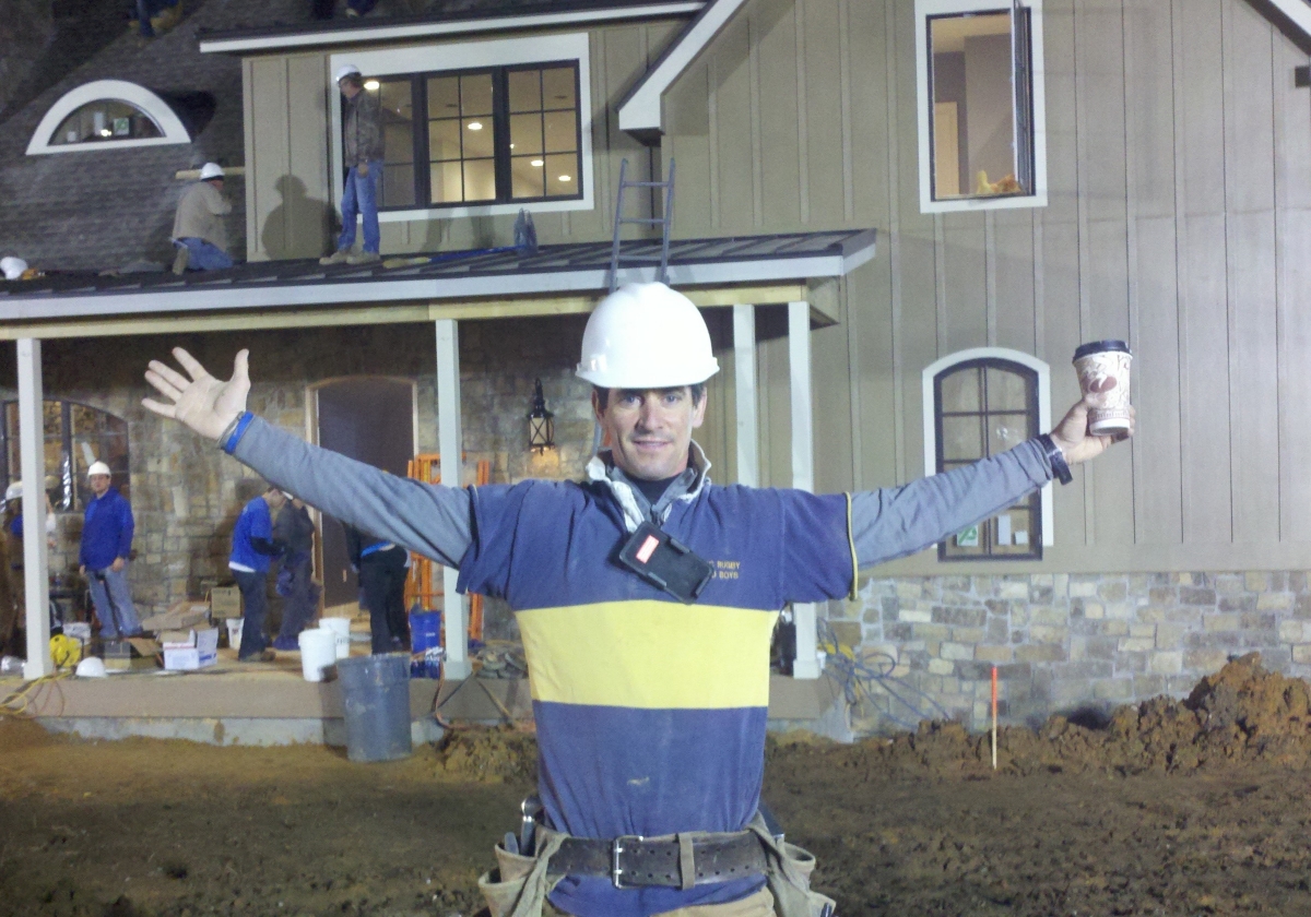 The project manager with a hard hat is standing in front of a home being renovated with his arms wide open.